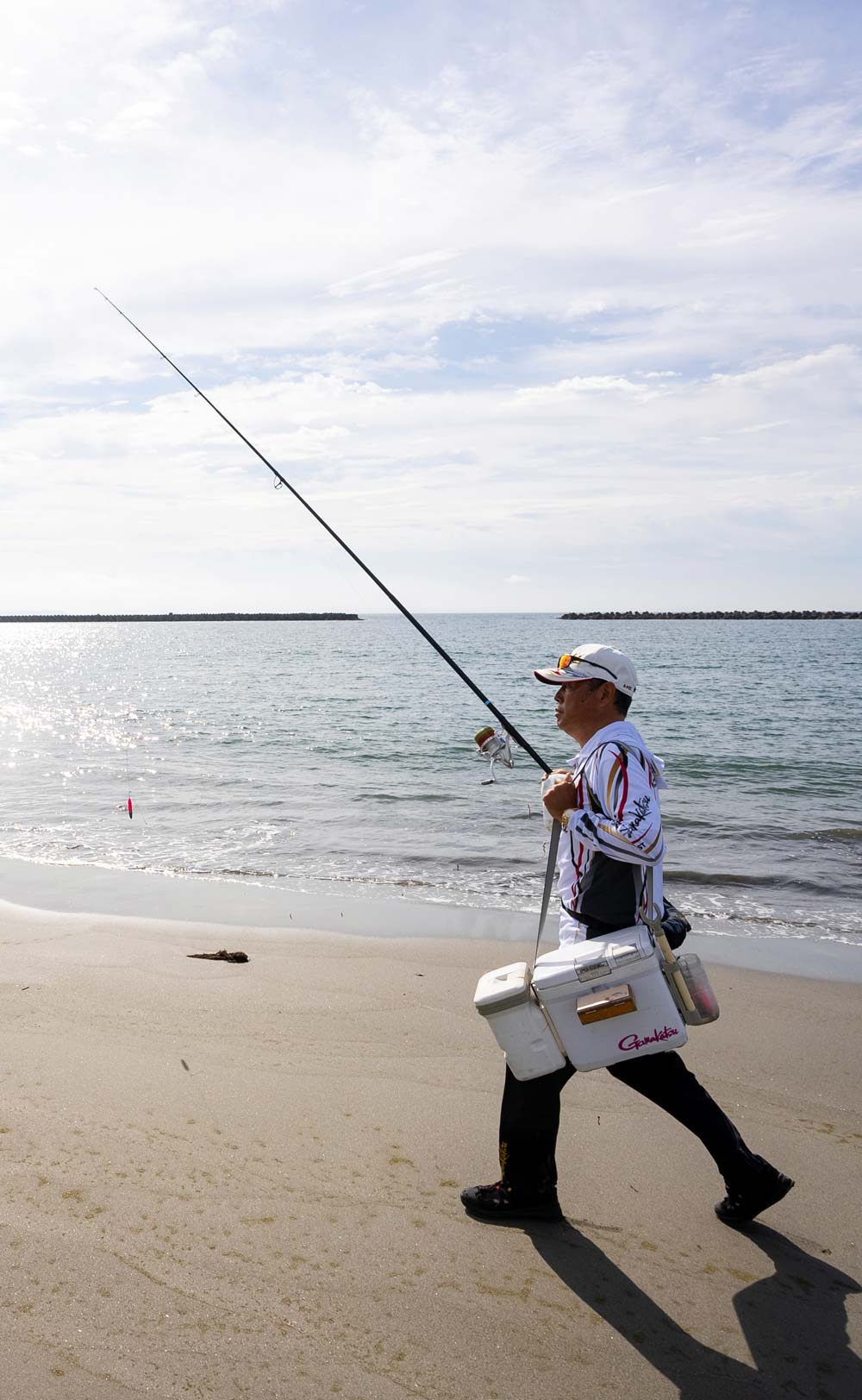 竿・クーラーボックスなどを持ち、砂浜を移動する矢野