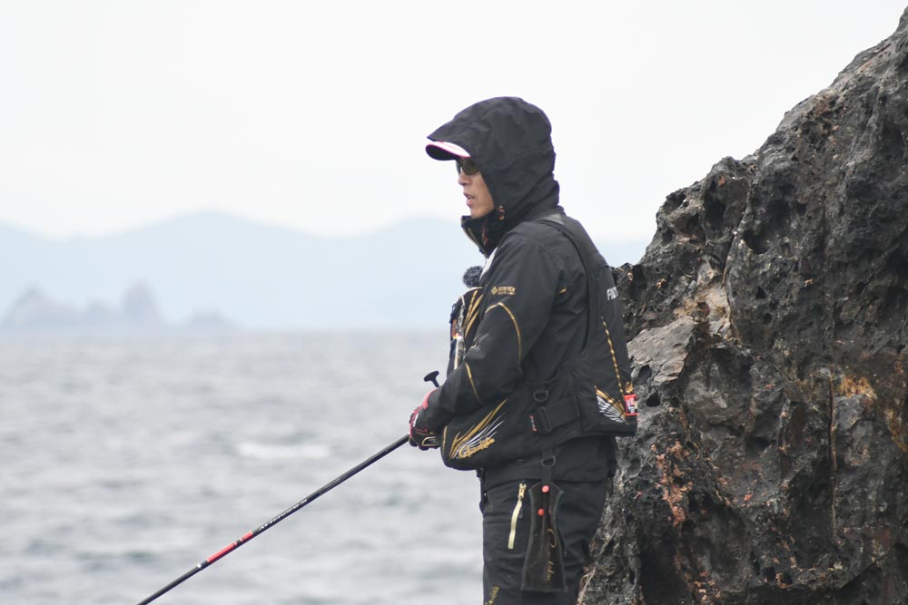 岩礁壁に立ち、海の様子を窺いながら竿を操作する敷地