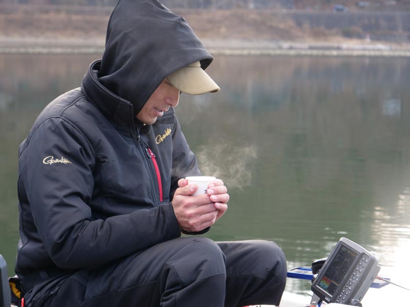 手漕ぎボート上で、湯気の上がるカップコーヒーを両手で包み持つ尾﨑