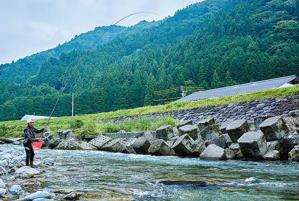 渓流の浅瀬に立ち、弧を描く竿を操作する廣岡