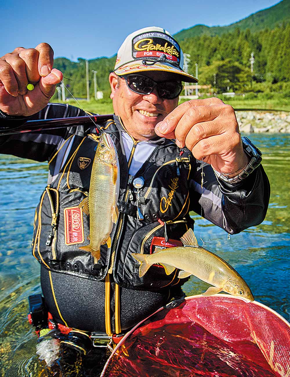 腰辺りまで浸かる深い川の中。釣り上げた鮎2尾がかかっている仕掛けを掲げ、満面の笑顔を見せる長谷川