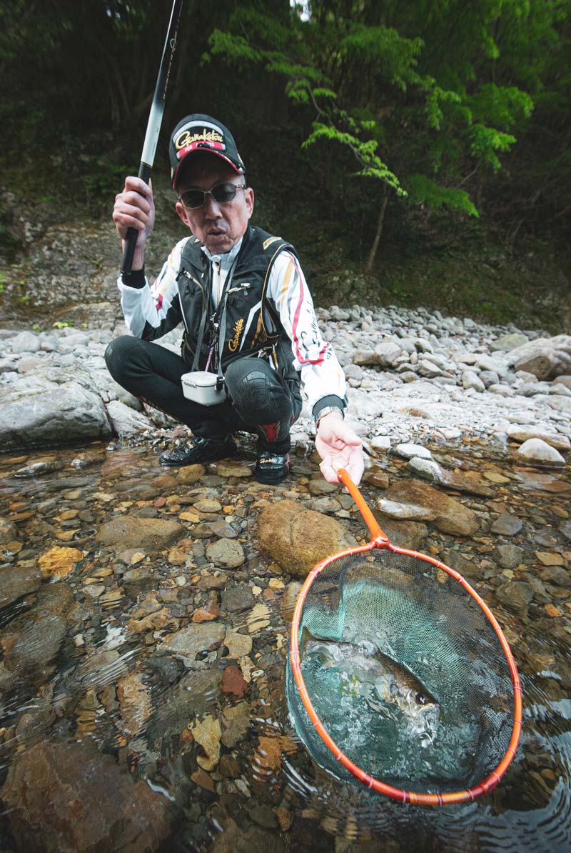 川岸にしゃがみ、網に収まる釣り上げた渓流魚を見せる廣岡