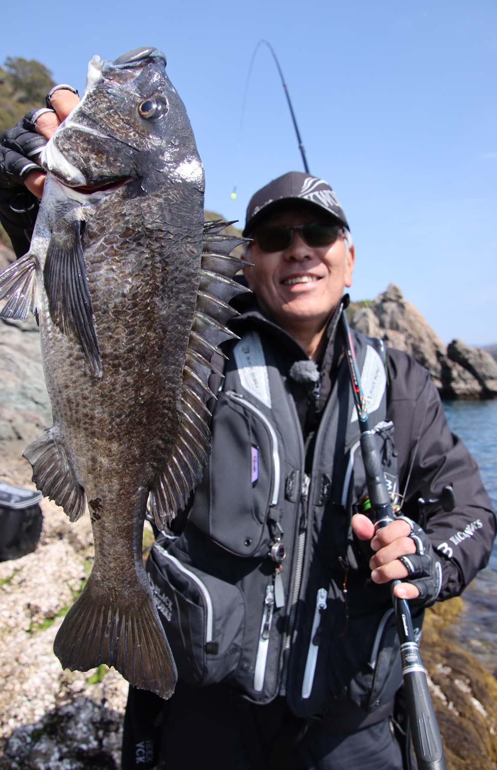 左手に持った竿を左肩に立てかけ、右手で釣り上げたチヌを掲げる中西