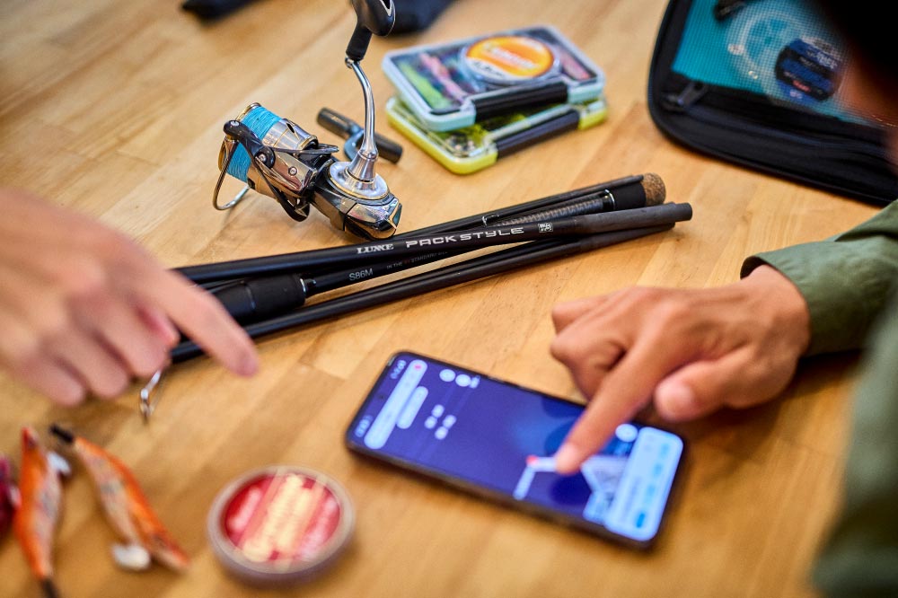 机の上に置かれたパックスタイルと釣り用品。その傍で、2人の人がスマートフォンの地図アプリを見ながら相談している。