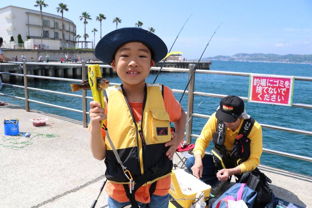 堤防（防波堤）を背に、フィッシュグリップで挟んだ魚を持って笑顔の男の子